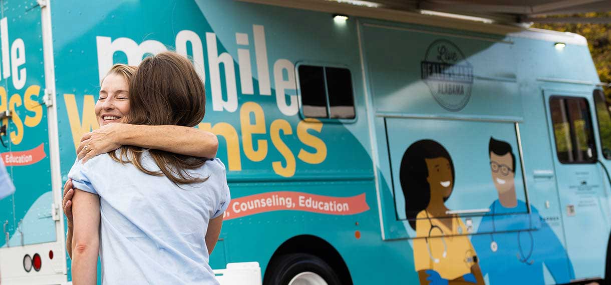 A blond woman hugs another in front of the van.