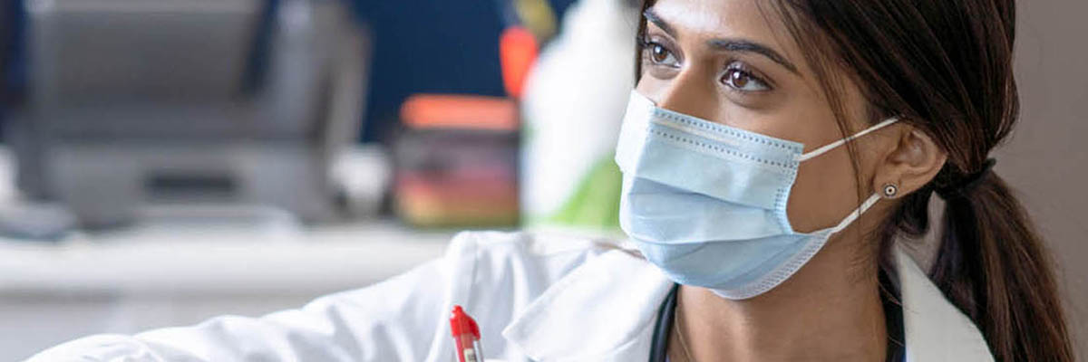 A female doctor bend's down to eye level as she talks with a cancer patient who is receiving her treatment intravenously. She is wearing a white lab coat and has a clipboard in hand as she takes notes. The patient is dressed casually and she sits back in the chair waiting for her treatment to finish, and both are wearing medical masks to protect them from COVID.