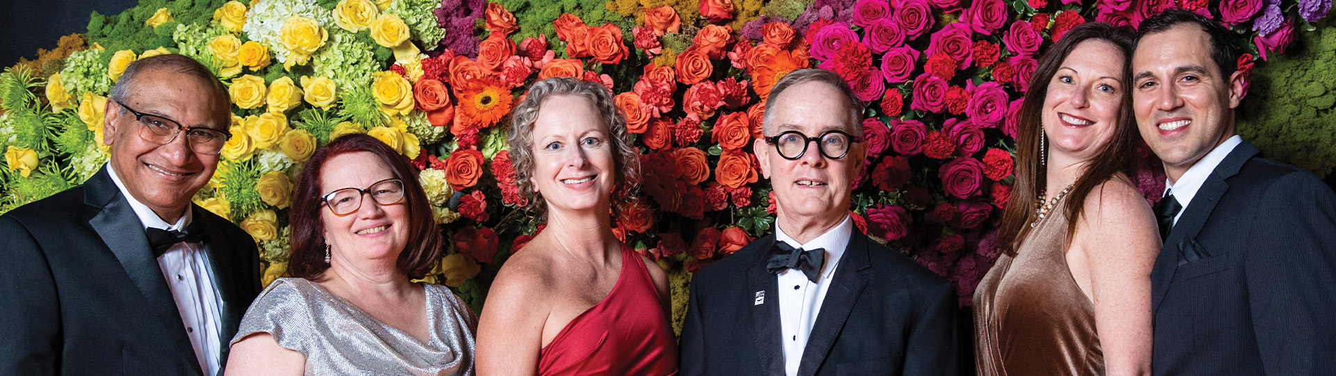 A group of attendees in formal wear pose in front of a multicolored floral wall.