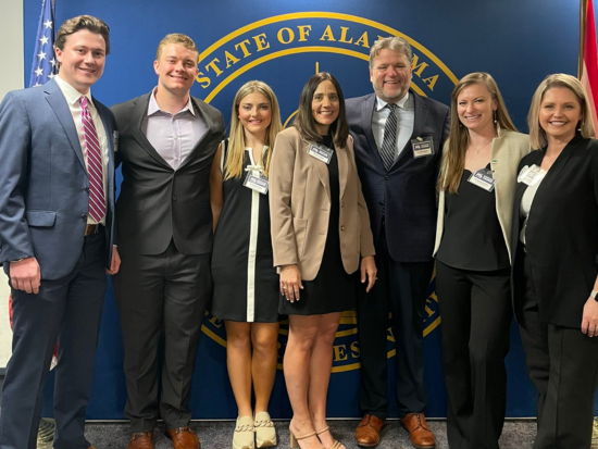 A group of seven professionally dressed individuals pose for a photo in front of a blue backdrop featuring the gold seal of the State of Alabama. They are standing close together, smiling at the camera, suggesting a formal event or professional gathering. An American flag is visible to the left, and a red flag is partially visible to the right.