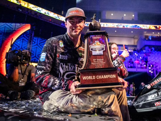 Easton Fothergill, wearing fishing gear and a cap, smiles while sitting on stage and holding the Ray Scott Trophy after winning the 55th Annual Bass Pro Shops Bassmaster Classic. The trophy reads "World Champion." Confetti covers the stage, and spectators and media are visible in the background.