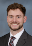 Professional headshot of a man wearing a dark suit jacket with a white shirt and a grey and red striped tie in front of a blue backdrop
