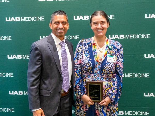 Two people smile while standing in front of a UAB Medicine background while holding a plaque