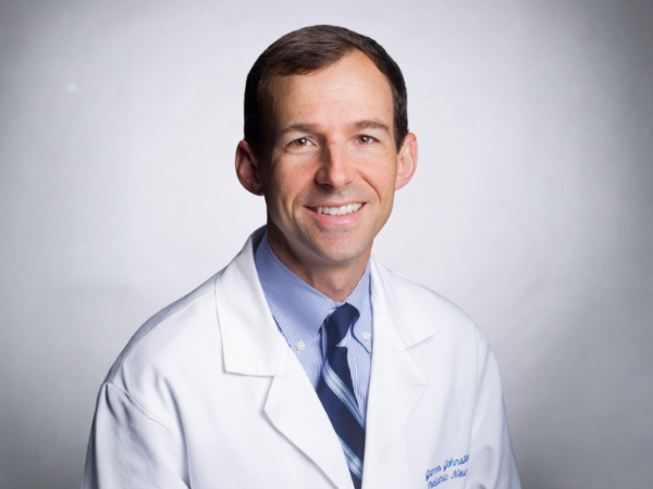 Professional headshot of a smiling man wearing a white medical coat, light blue dress shirt, and blue striped tie against a light gray background.