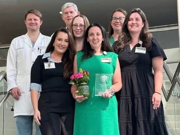 Group of 7 individuals where woman center is wearing a green dress holding a bouquet and a glass award.