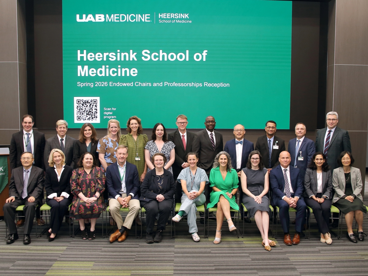 Group of faculty members sitting and standing in front of a screen with a green background and white text that reads Heersink School of Medicine Spring 2026 Endowed Chairs and Professorships Reception