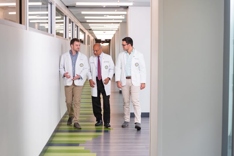 Dr. Anupam Agarwal (Senior Vice President/Dean, Heersink School of Medicine) is mentoring medical students, all wearing white medical coats, while walking in an interior hallway.