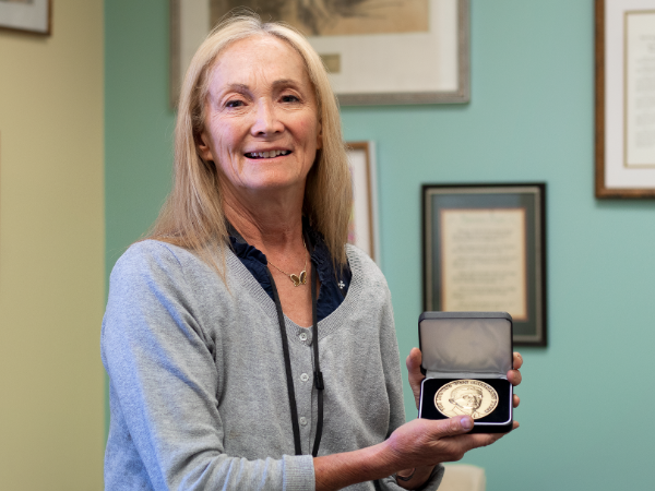 Person with long blonde hair holds an open presentation case displaying a gold medal in an office.
