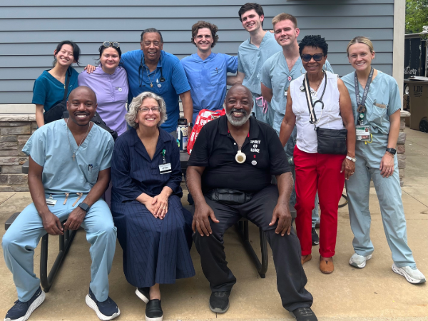 Group photo of 10 health care professionals standing and seated outdoors in front of a gray-sided building. Three people are seated in the front row, with seven standing behind them. Several individuals are wearing scrubs or medical attire, and some have stethoscopes or ID badges visible. The group is gathered on a concrete patio area for a posed photo.