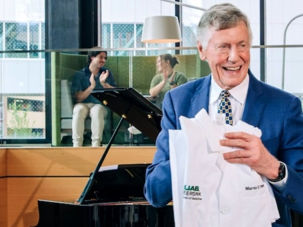 Dr. Heersink smiles while holding new medical white coat in front of grand piano.