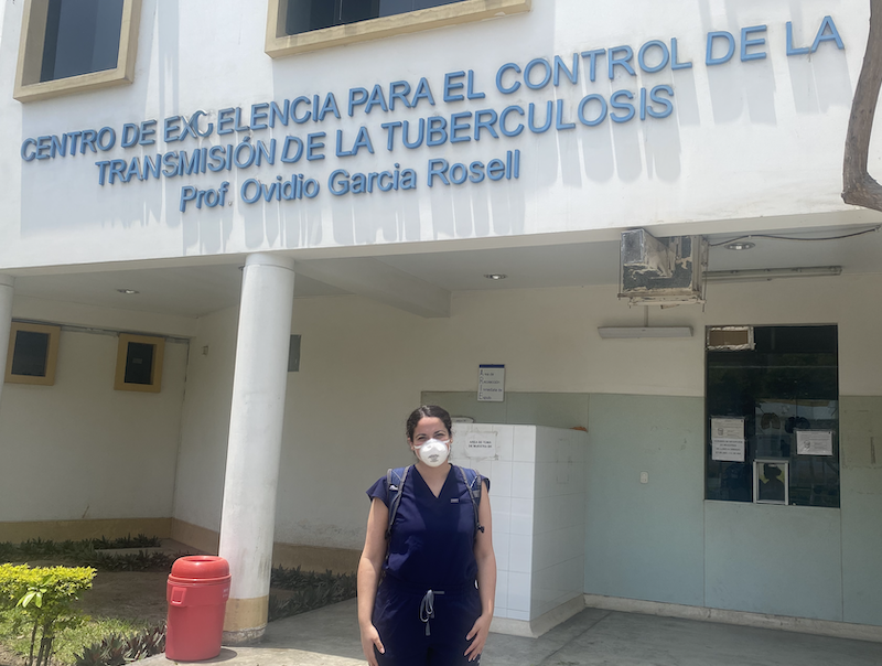 Proenza stands in front of a Socios En Salud TB clinic in Peru with a mask and scrubs on