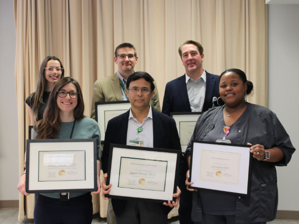 Group of six employees standing indoors and holding framed award certificates during a recognition event.