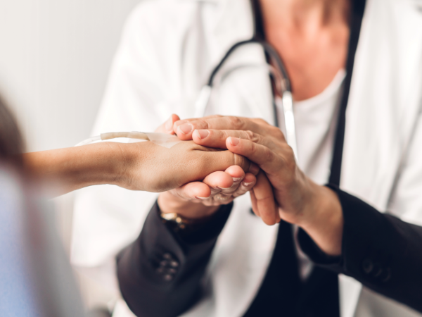 Close-up of a healthcare professional in a white coat with a stethoscope gently holding a patient's hand during a clinical visit. The image is softly focused, emphasizing compassionate care and support.