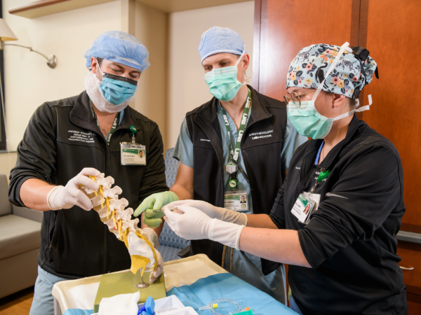 Three UAB anesthesiology residents wearing surgical masks, caps, and gloves stand around a medical table, collaboratively examining a spine model and demonstrating a procedure using medical tools. A draped tray with supplies is in the foreground, and cabinets and a countertop are visible in the background.