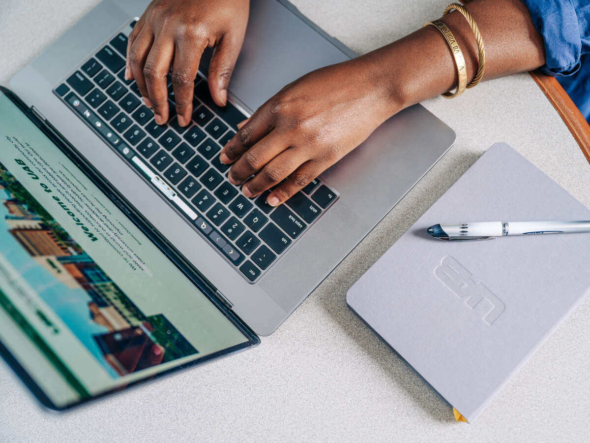 An individual types on a laptop with a UAB-branded notebook on the table.