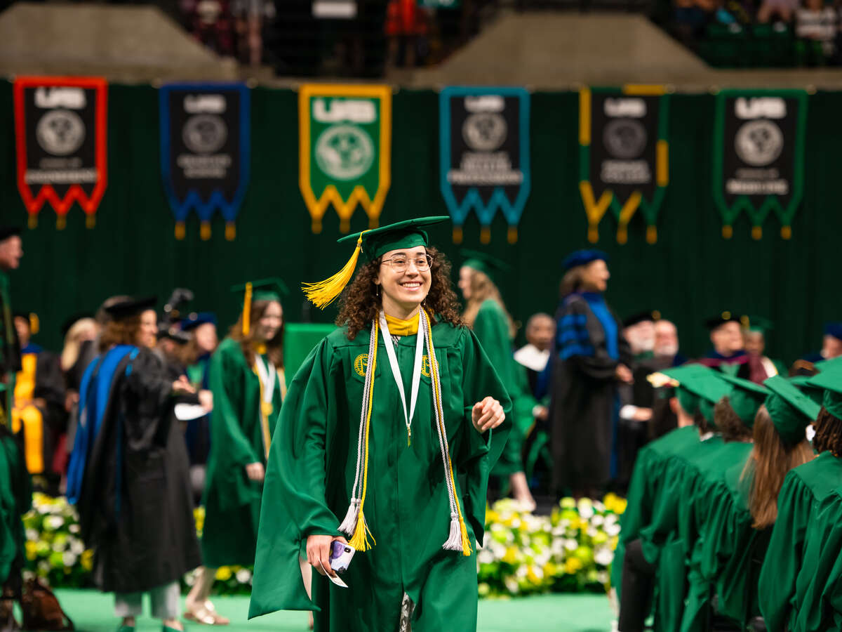 Smiling female graduating student is wearing green cap and gown and walking from stage back to seat during the ceremony in Bartow Arena, April 2024.