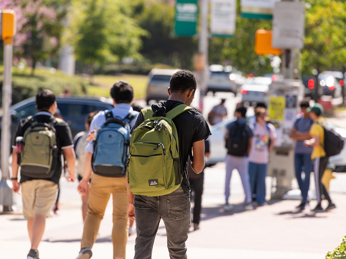 Back, students are wearing backpacks and walking on sidewalk along University Boulevard with other students standing beside a crosswalk near road traffic in the background, August 2024.