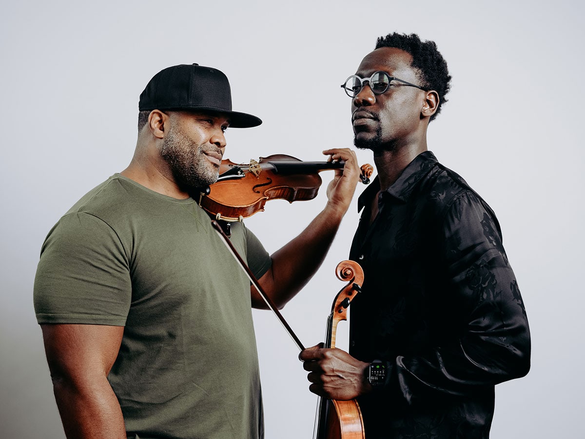 Two men, dressed in casual clothing with one wearing a baseball cap, hold string instruments.