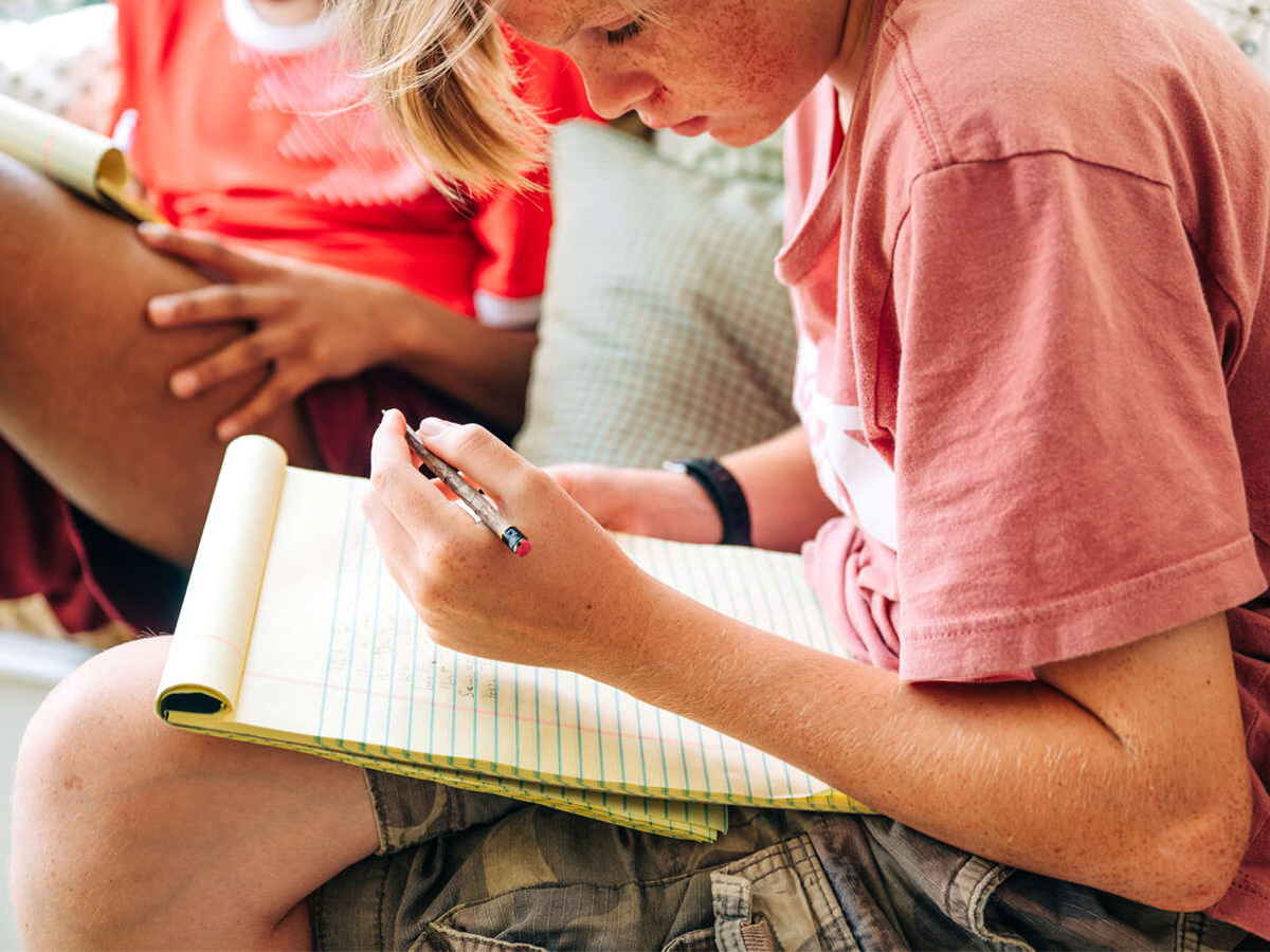 From side, a male teen student is writing on a notepad in his lap.