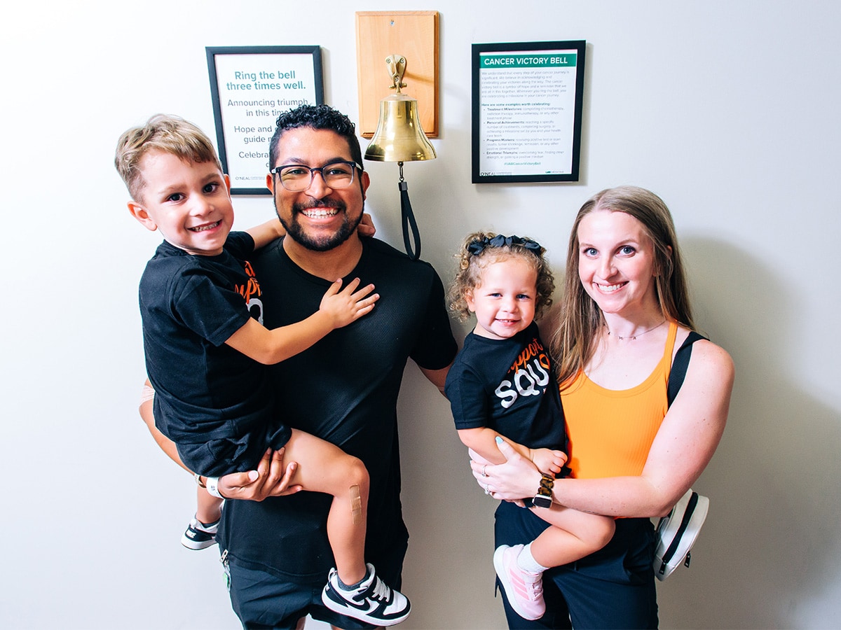 Photo of a mom and dad holding their two kids in front of a bell hanging on a wall. 