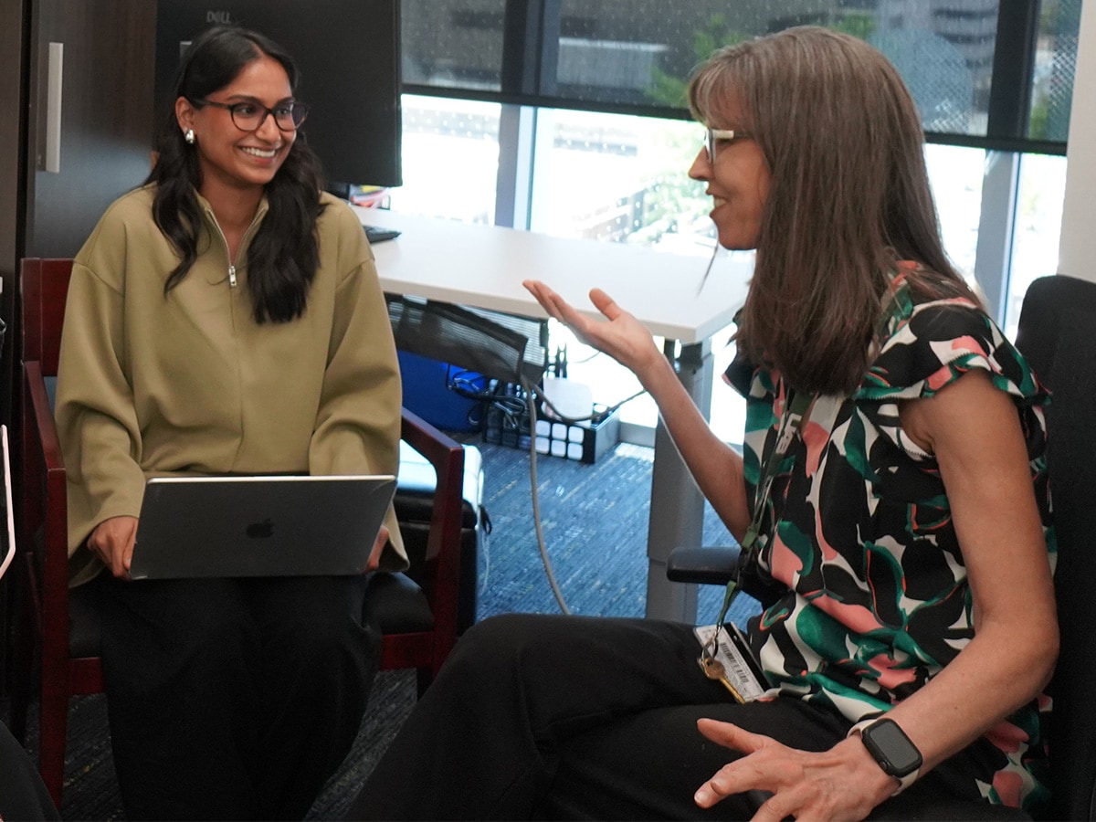 Image of Dua Khan wearing a green zip up blouse with a laptop in her lap in a office setting talk to a woman. 