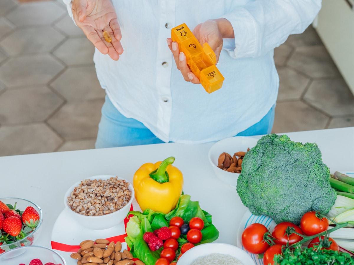 Image shows the lower half of a middle-aged woman holding a supplement and pill organizer, with healthy foods spread on the counter below her, including strawberries, bell peppers, broccoli, and tomatoes.
