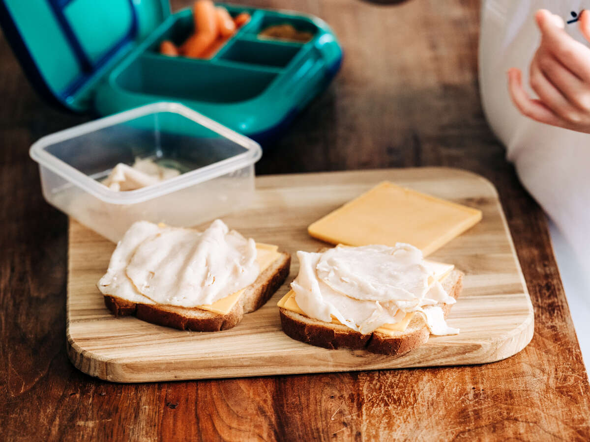 From side, close-up of boy's hands making a deli sandwich on a cutting board while standing beside partially packed lunch box. 