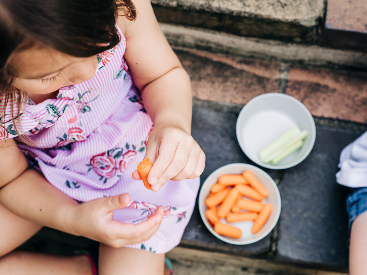 Shot from above, toddler girl is holding a baby carrot while sitting on outdoor steps beside small bowls of baby carrots and pieces of celery.