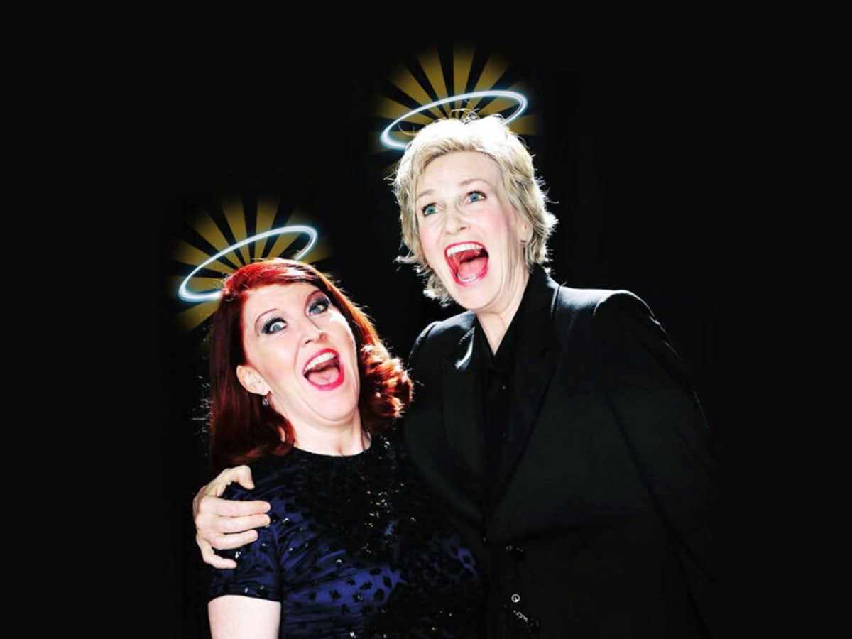 The two performers, dressed in blue and black and standing in front of a black background., side hug each other while singing. Angel halos shine above their heads.