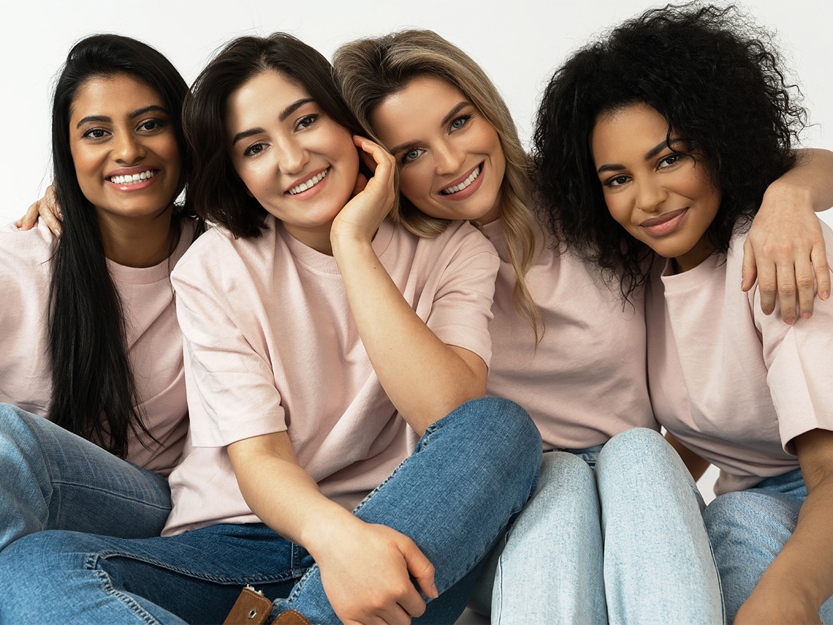 Group of four women from various racial backgrounds sitting close together wearing light pink t-shirts and blue jeans.
