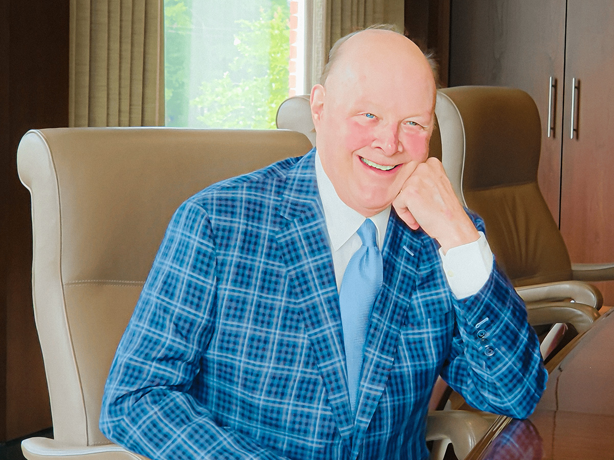 Photo of Norman Estes sitting in a tan desk chair at a brown conference table in a blue pattern sports coat and light blue tie. 