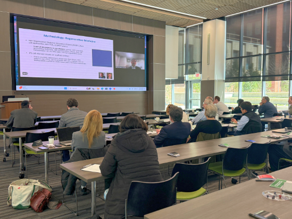 Vision Science Research Center members gather in a modern lecture space during the VSRC retreat, where attendees watch a keynote presentation delivered virtually. The session features slides projected on a large screen, with researchers seated at long tables, engaging in discussion and learning about current topics in vision science and research priorities.