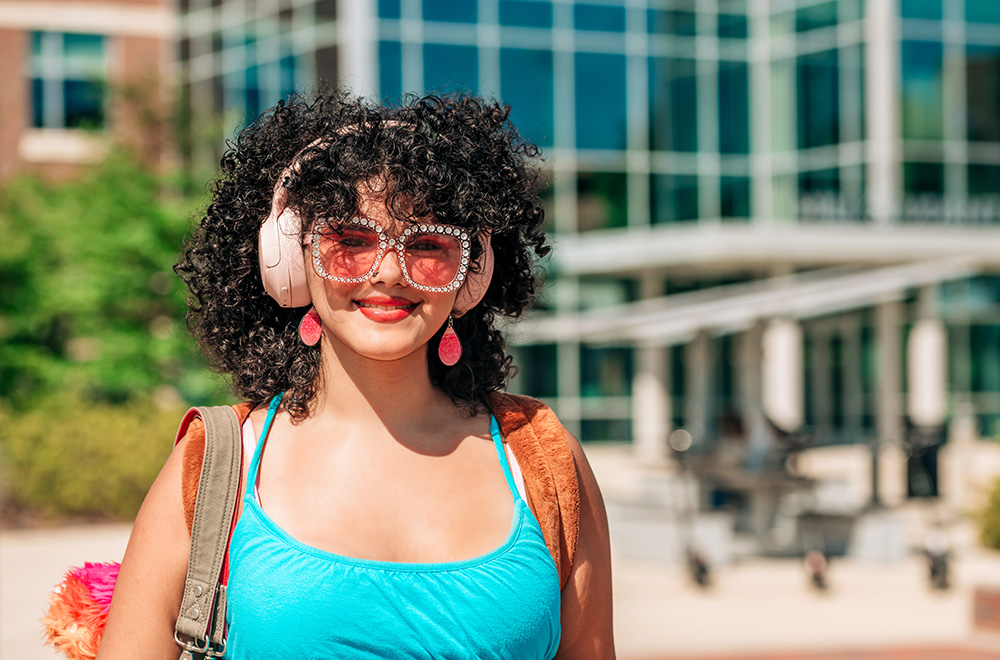 A student wearing sunglasses and headphones smiles at the camera outside of the Hill Student Center on UAB's campus