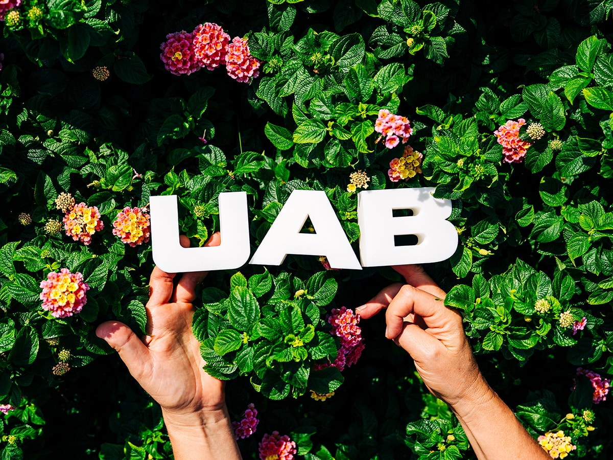 Hands hold 3D printed letters spelling U-A-B in front of flowers on the Campus Green