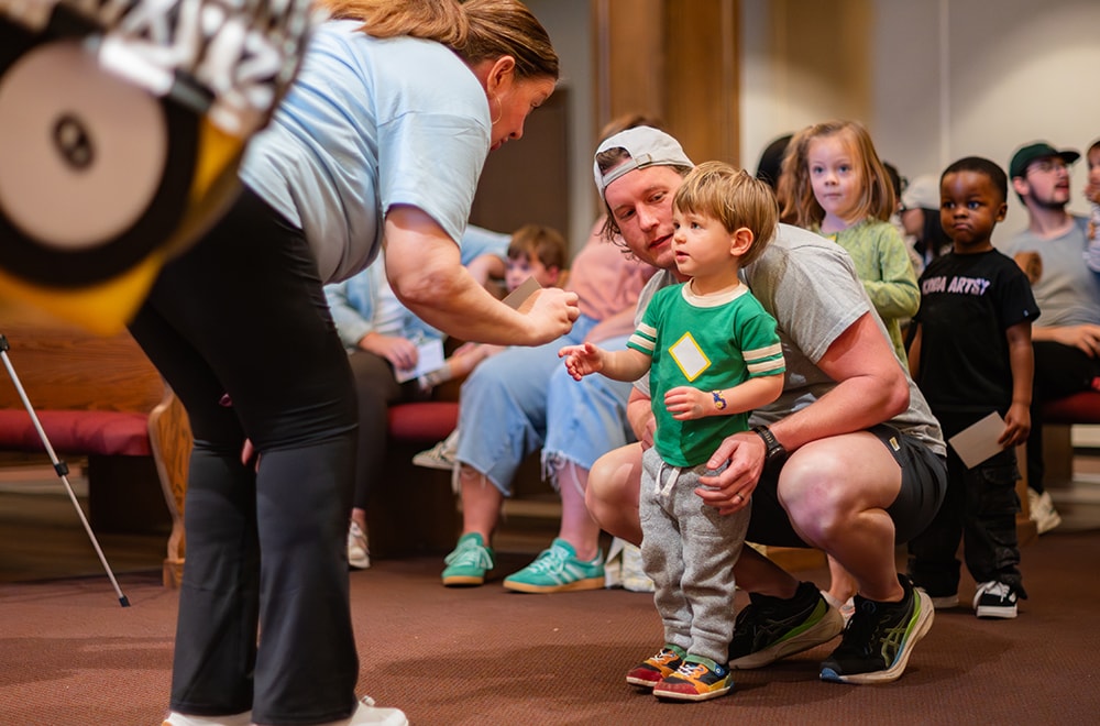 A small child and his father are talking to a facilitator, who is bending down to listen, while a crowd of kids gathers behind in the audience