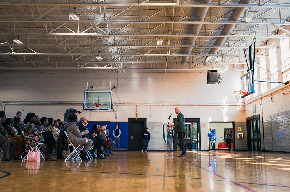 UAB President Ray Watts speaks at a podium in front of an audience in a school gymnasium.