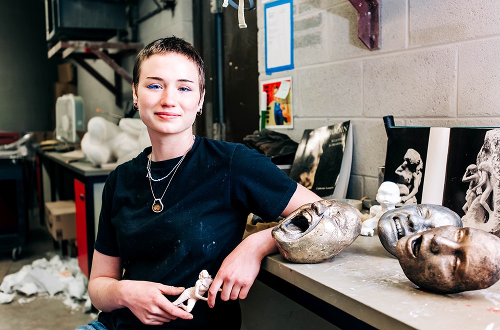 A student, wearing a necklace and blue shirt, poses in a UAB studio with her sculptures of emotion-laden faces on a table next to her, and photographs of Rodin sculptures leaned against the wall behind