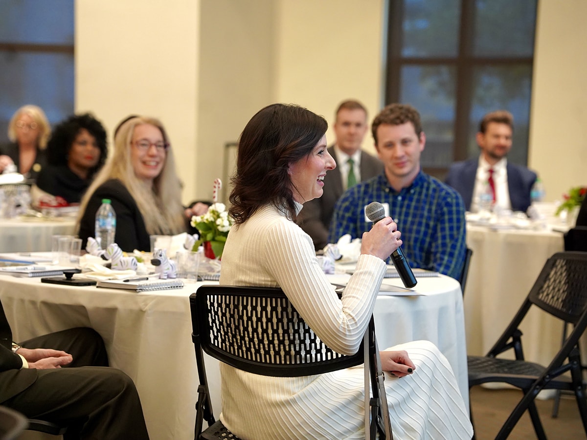 A local elected official, seated, speaks at the microphone while the room full of attendees looks on