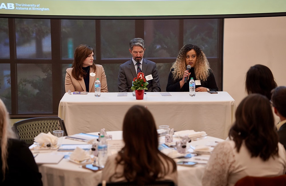 Three panelists sit at a podium in front of a large room, with one talking in a microphone. Audience members at tables with their backs to the camera listen