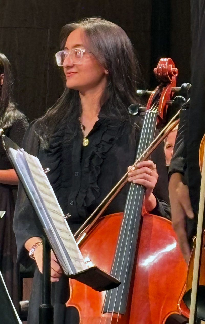 Tamana Patel stands holding her cello, wearing all black. A music stand and other people are visible in this performance photo.