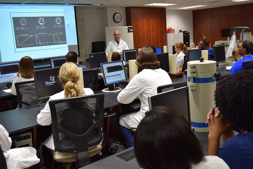 Students in white medical coats sit at tables in a classroom, watching their teacher at a large digital display. 