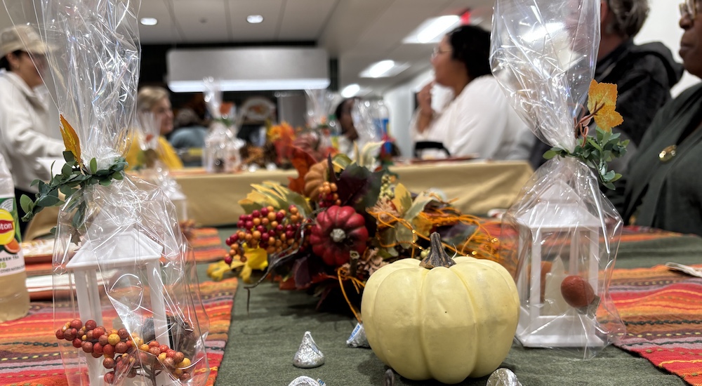 Thanksgiving table with people talking and eating in the background. The foreground has Thanksgiving decor of small pumpkin and cornucopia.