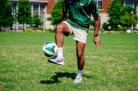 Close-up, legs of male student kicking a soccer ball on the Campus Green, June 2019.