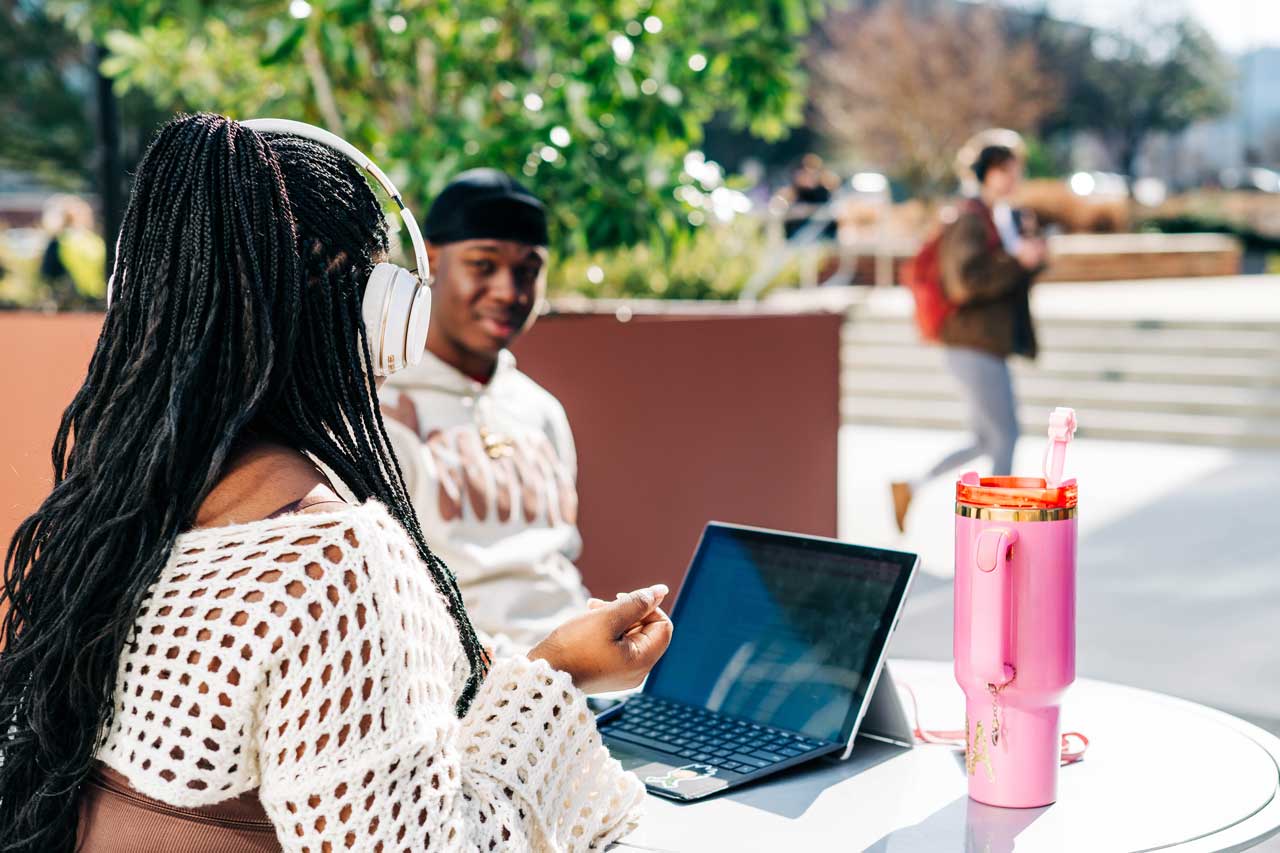 Student sits outdoors with a laptop while talking with another student at a table.