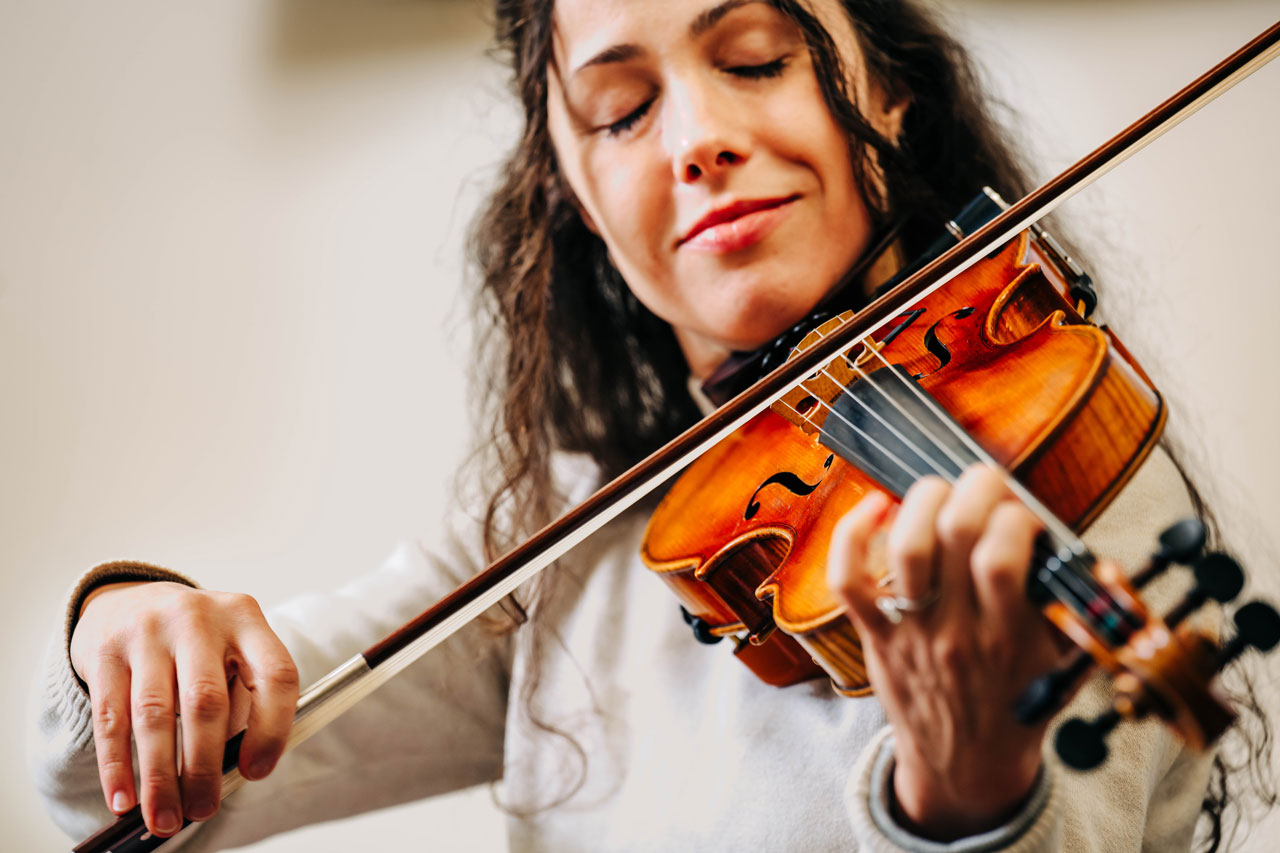Person plays a violin indoors, holding the instrument under her chin while drawing the bow across the strings.