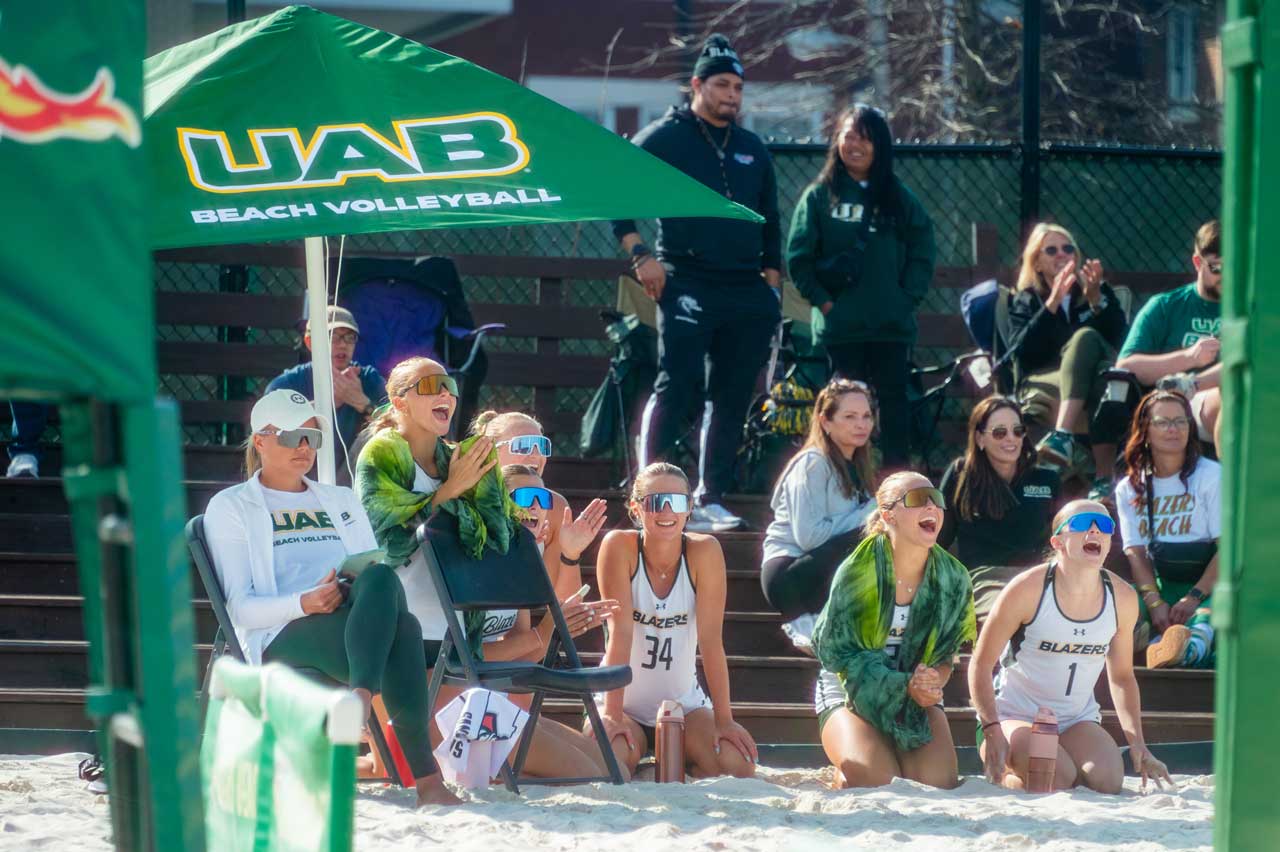 Several beach volleyball players and fans are cheering the team at the UAB Beach Volleyball Courts.