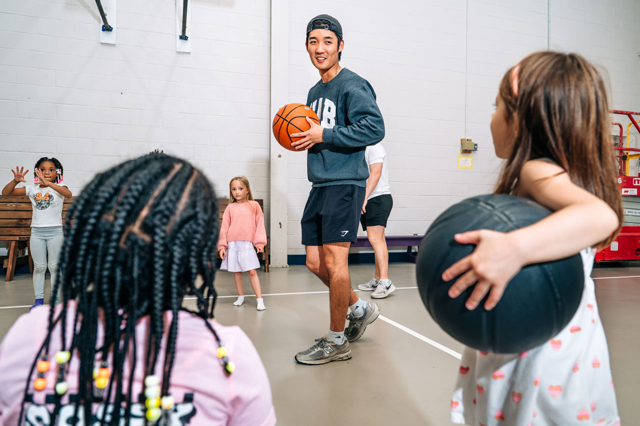 Coach leads children holding basketballs during a gym activity.